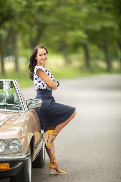 Mooie vrouw in een rok en hoge hakken leunt met haar rug tegen een vintage cabriolet geparkeerd langs de weg.