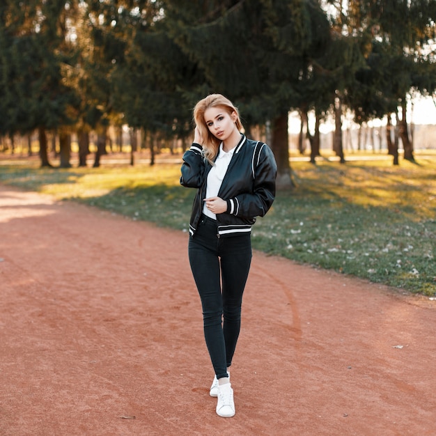 Mooie vrouw in een zwarte jas, zwarte spijkerbroek en witte schoenen in het park op een zonnige dag