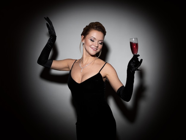 Mooie vrouw in een zwarte jurk met een halslijn en lange fluwelen handschoenen die dansen en champagne vasthouden...