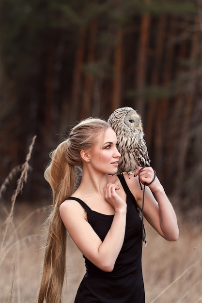 Mooie vrouw in een zwarte jurk met een uil op zijn arm. Blonde met lang haar in de natuur met een uil. Romantisch delicaat beeld van een meisje