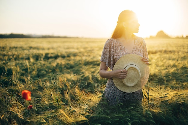 Mooie vrouw in gebloemde jurk staande in gerstveld bij zonsondergang licht Atmosferisch rustig moment rustiek langzaam leven Stijlvolle vrouw strohoed vasthouden en genieten van avond zomer platteland