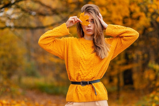 Mooie vrouw in gebreide gele trui bedekt haar gezicht met een geel herfstblad buiten in het bos met gouden bladeren