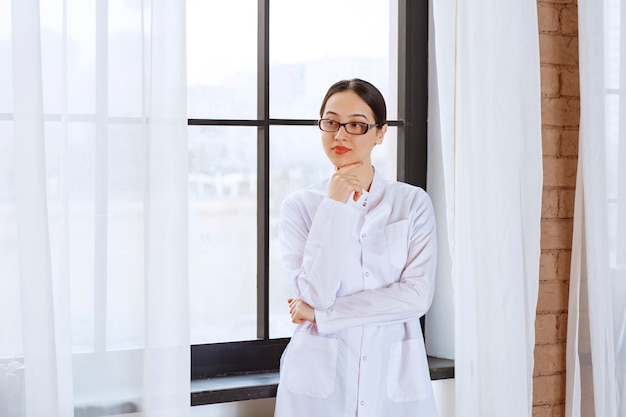 Mooie vrouw in glazen en laboratoriumjas die zich dichtbij venster bevindt.