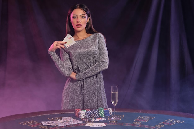 Mooie vrouw in grijze jurk toont twee kaarten terwijl ze poseren aan speeltafel in casino Zwarte rook achtergrond met kleurrijke achtergrondverlichting Gokken entertainment poker champagne close-up