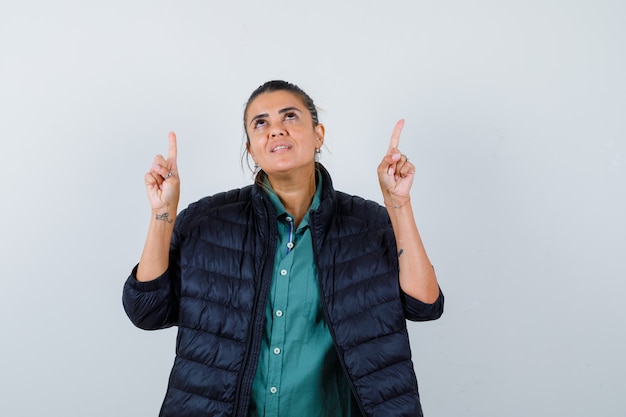 Mooie vrouw in groen shirt, zwarte jas die met wijsvingers omhoog wijst, naar boven kijkt en er gelukkig uitziet, vooraanzicht.