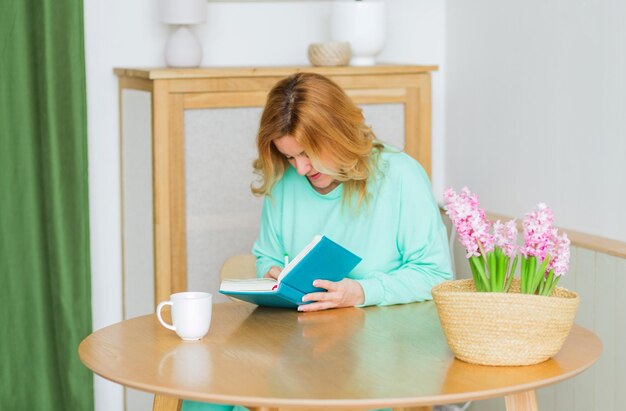 Mooie vrouw in groene pyjama zit aan de keukentafel met een notitieboekje in haar handen.