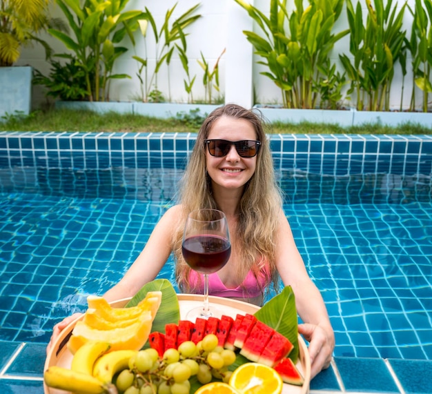 Foto mooie vrouw in het zwembad met een glas wijn en een drijvende dienblad met fruit.