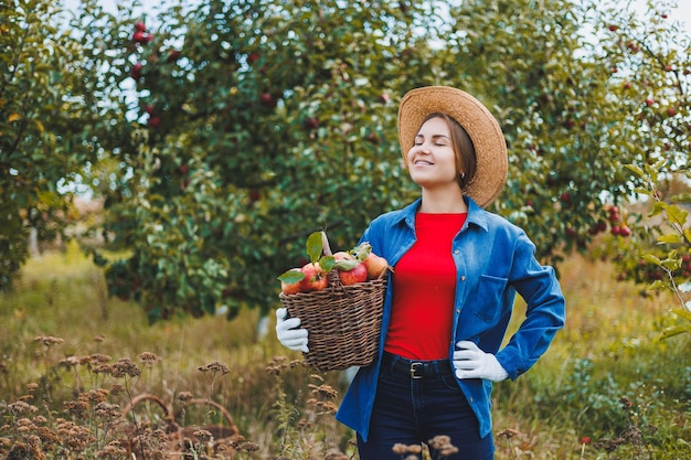 Mooie vrouw in hoed en shirt in herfsttuin met rijpe appels in mand en glimlachen Een vrouw verzamelt rijpe appels Appels oogsten in de herfst