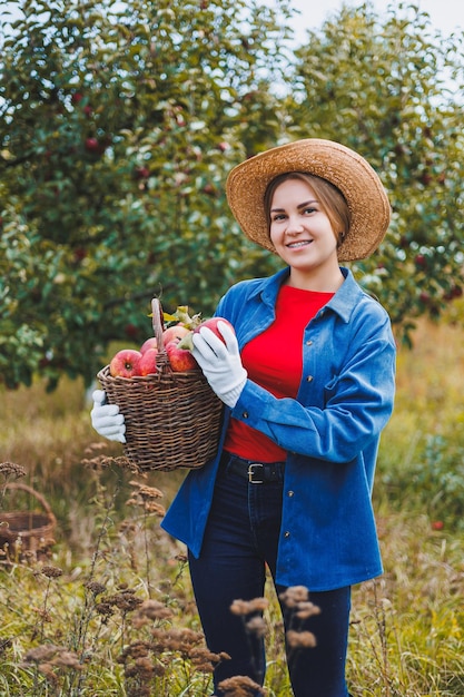 Mooie vrouw in hoed en shirt in herfsttuin met rijpe appels in mand en glimlachen Een vrouw verzamelt rijpe appels Appels oogsten in de herfst