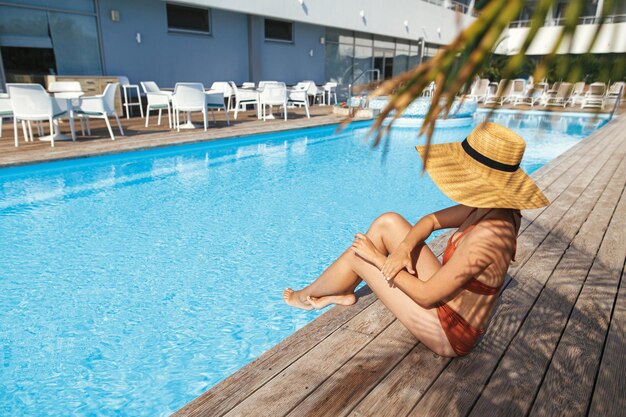 Mooie vrouw in hoed ontspannen op houten pier bij blauw zwembad genieten van zomervakantie in het resort