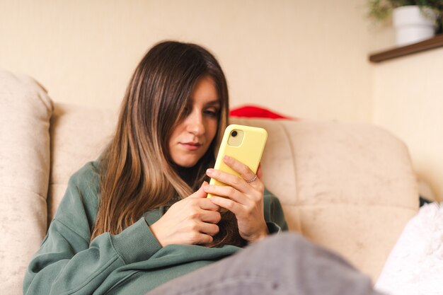 Mooie vrouw in hoody sms'en en chatten op de mobiele telefoon, ontspannen vanuit huis, zittend op de bank.