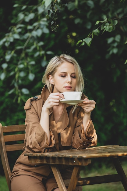 Mooie vrouw in jas met kopje koffie aan houten tafel in een tuin met bomen op de achtergrond