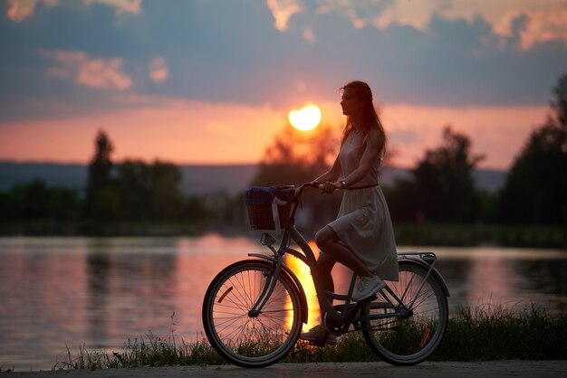 Mooie vrouw in jurk rijdt op een vintage fiets met mand op weg in de buurt van de rivier bij avondrood met wolken