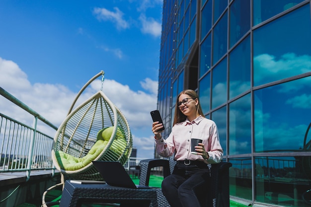 Mooie vrouw in klassieke bril en trendy outfit die een bericht leest op een mobiele telefoon die naast een modern glazen gebouw staat