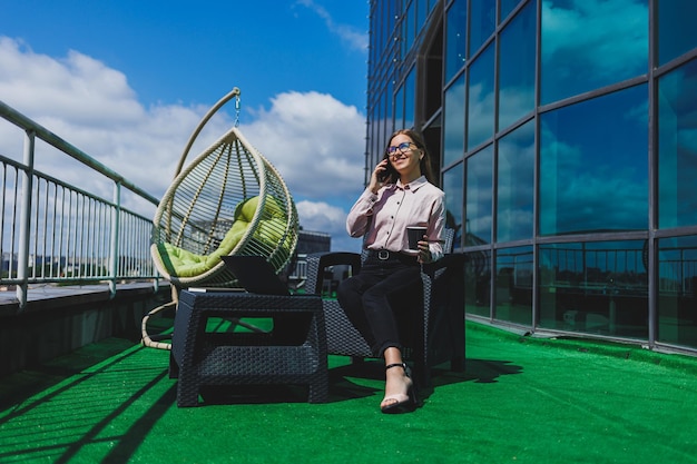 Mooie vrouw in klassieke bril en trendy outfit die een bericht leest op een mobiele telefoon die naast een modern glazen gebouw staat