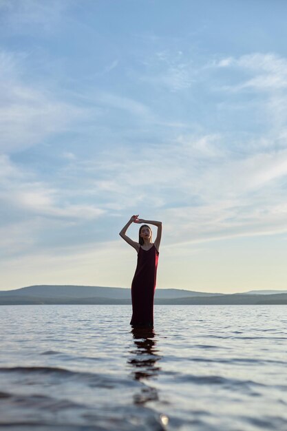 Mooie vrouw in lichte rode jurk staat in het water van de zee van het meer bij zonsondergang. Weerspiegeling van het meisje en de lucht in het water