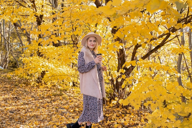 Mooie vrouw in modieuze kleding en hoed drinkt koffie in de natuur in de herfst.