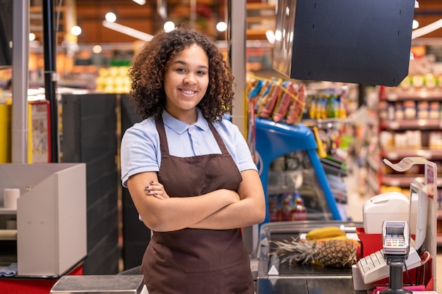 Mooie vrouw in schort permanent door kassa in supermarkt en kruising armen door borst op muur van planken met voedingsproducten