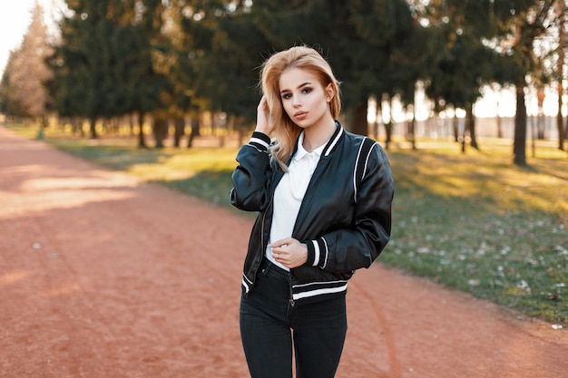 Mooie vrouw in stijlvolle zwarte jas en spijkerbroek wandelen in het park op een zonnige dag