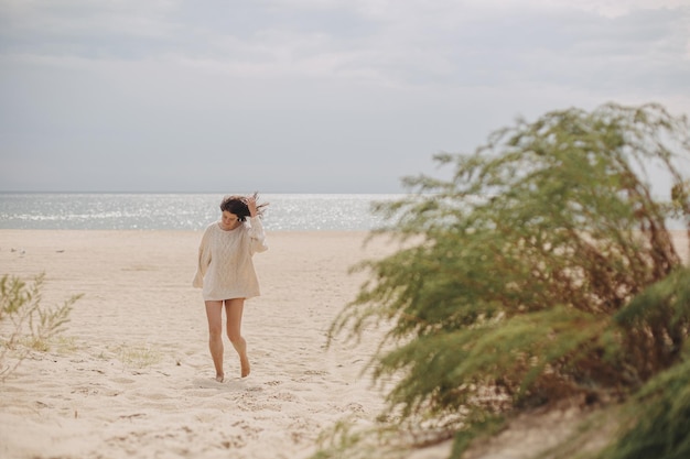 Mooie vrouw in trui met winderig haar lopen op zandstrand bij groen gras en zee vakantie