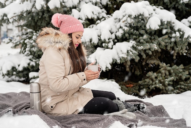 Mooie vrouw in warme winterkleren met beker die hete thee of koffie buiten drinkt op een besneeuwde dag