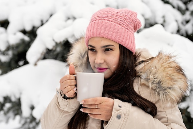 Mooie vrouw in warme winterkleren met beker die hete thee of koffie buiten drinkt op een besneeuwde dag