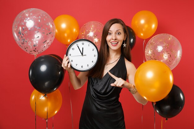 Mooie vrouw in zwarte jurk vieren wijzende wijsvinger op ronde klok in handen op heldere rode achtergrond luchtballonnen. St. Valentijnsdag, Gelukkig Nieuwjaar, verjaardag mockup vakantie feest concept.