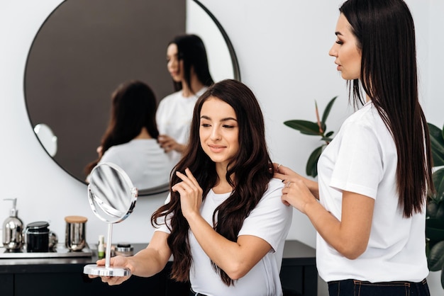 Mooie vrouw Kapper maakt haarsnit op een schattige vrouw cliënt in een kapsalon binnenshuis Plezier dame kijken naar de hand spiegel