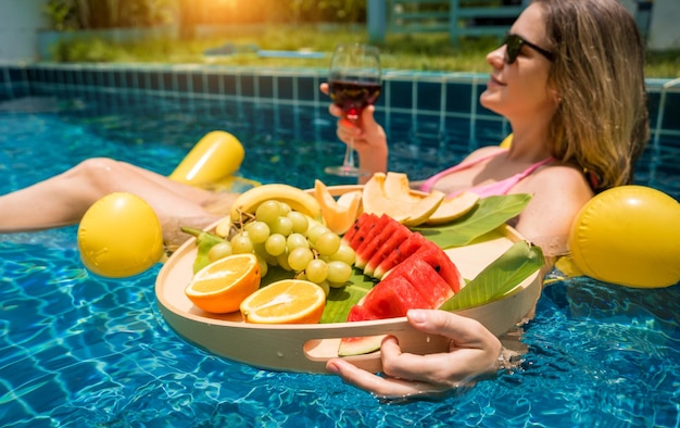 Mooie vrouw ligt op een drijvende hangmat in het zwembad met wijn en een dienblad met fruit