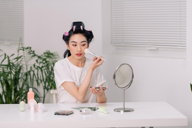 Mooie vrouw maakt blos op haar gezicht met make-upborstel terwijl ze thuis in een kamer aan een tafel zit en in de spiegel kijkt