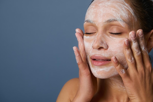 Mooie vrouw masseert haar gezicht terwijl ze make-up verwijdert met een schuimreinigend cosmetisch product, en haar huid verfrist met een exfoliant schoonheidsproduct, geïsoleerd over grijze achtergrond. Detailopname.