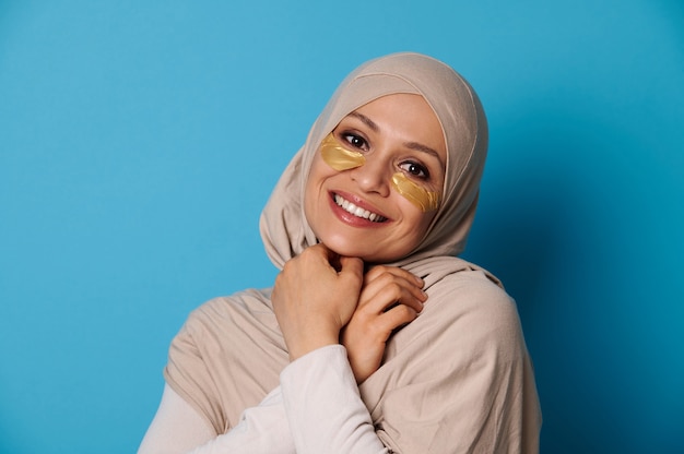 Mooie vrouw met bedekt hoofd in hijab, glimlachend in de camera met hydrogel collageen patches onder de ogen. Geïsoleerd