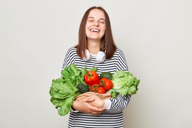 Mooie vrouw met bruin haar met een gestreept overhemd dat geïsoleerd over een grijze achtergrond staat en verse groenten koopt voor een gezond dieet, lachend geniet