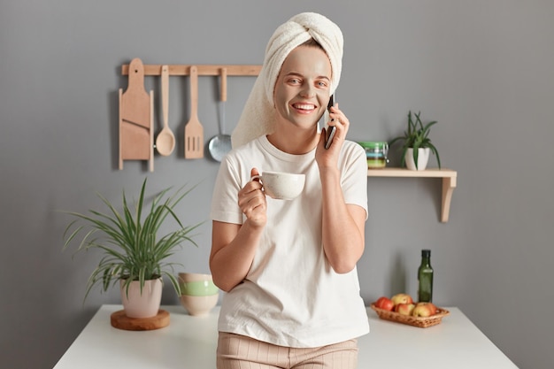 Mooie vrouw met cosmetisch masker op gezicht 's ochtends koffie drinken en praten via smartphone thuis in de keuken jonge vrouw gewikkeld in een handdoek die de oproep telefonisch beantwoordt tijdens de huidverzorgingsbehandeling