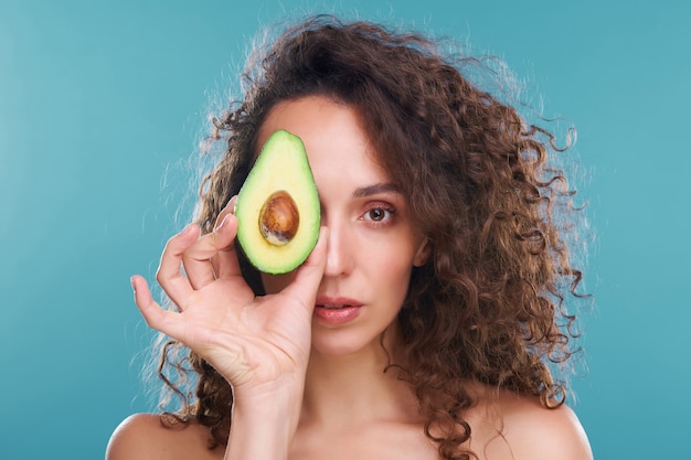 Mooie vrouw met een gezonde huid en luxueus golvend haar die naar je kijkt met het linkeroog terwijl ze de helft van de verse avocado vasthoudt aan een andere