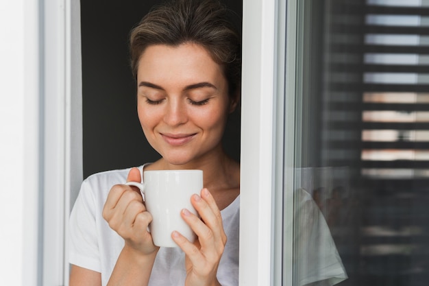 Mooie vrouw met een kop warme koffie die door het raam kijkt