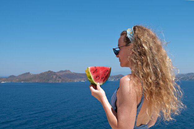 Mooie vrouw met een plakje watermeloen die zwembroek draagt ?? die rode rijpe watermeloenplak met de vakantiezomer van het overzees bekijken.