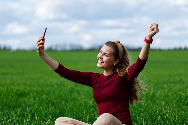 Mooie vrouw met een telefoon in haar handen zit op het gras. Het meisje kijkt naar zichzelf in de camera van de telefoon en maakt selfie-foto's. Ze lacht en geniet van een warme dag. Conceptfoto op smartphone