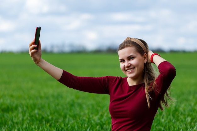 Mooie vrouw met een telefoon in haar handen zit op het gras. Het meisje kijkt naar zichzelf in de camera van de telefoon en maakt selfie-foto's. Ze lacht en geniet van een warme dag. Conceptfoto op smartphone