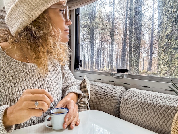 Mooie vrouw met een wollen hoed kijkt uit het raam van de camper. Vrouw geniet van het bos vanuit haar camper.