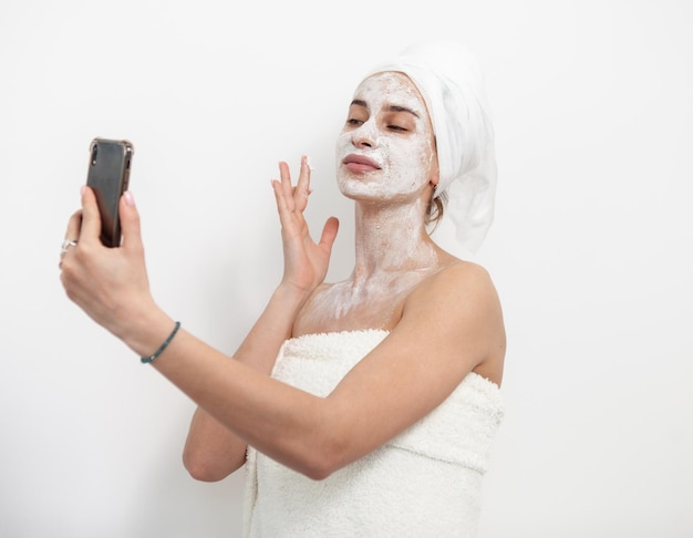 Mooie vrouw met gezichtsmasker kijkt naar scherm van smartphone op witte achtergrond Huidverzorgingsconcept