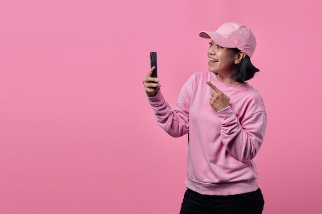 Mooie vrouw met hoed glimlachend gelukkig met handgebaar wijzend op smartphone op roze achtergrond