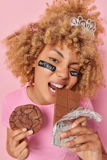 Mooie vrouw met krullend haar bijt reep chocolade houdt heerlijk koekje probeert niet te huilen dus eet snoep heeft verslaving aan suiker terloops gekleed geïsoleerd over roze achtergrond Lekker dessert