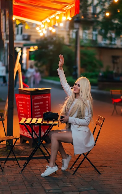 mooie vrouw met lang blond haar in een lichte jurk en jas zit in een straatcafé