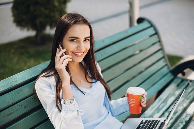 Mooie vrouw met lang donker haar die direct lachend kijkt terwijl ze aan de telefoon praat met een kopje koffie in de ene hand en met een laptop op haar benen buiten.