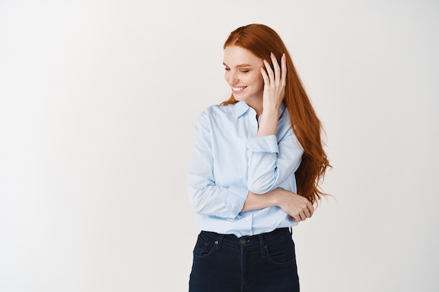 Mooie vrouw met lang rood haar dat bloost en verlegen wegkijkt, over een witte muur staat en glimlacht