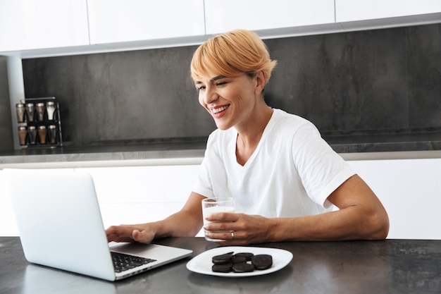 Mooie vrouw met laptopcomputer zittend aan de keukentafel, melk drinken uit een glas, koekjes eten