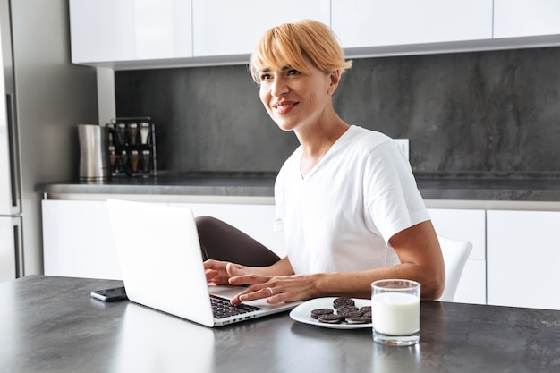 Mooie vrouw met laptopcomputer zittend aan de keukentafel, melk drinken uit een glas, koekjes eten