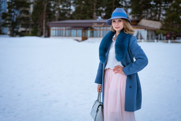 Mooie vrouw met make-up, in een blauwe jas en hoed, poseren in de natuur in de sneeuw