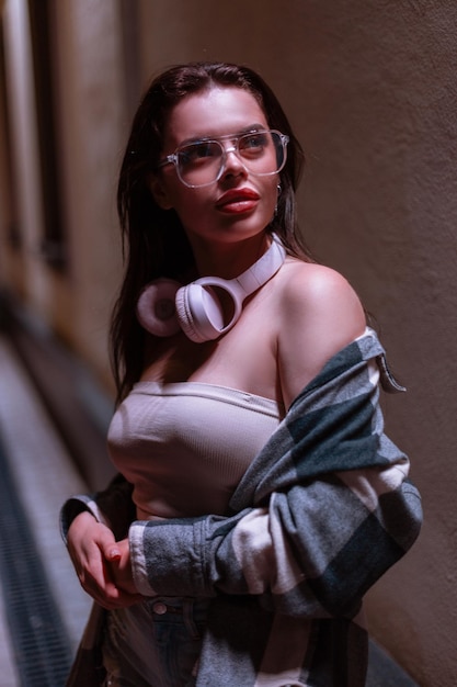 Mooie vrouw met modieuze brillen en roze koptelefoon in stijlvolle top en geruite hemd loopt in de nachtstad bij een muur